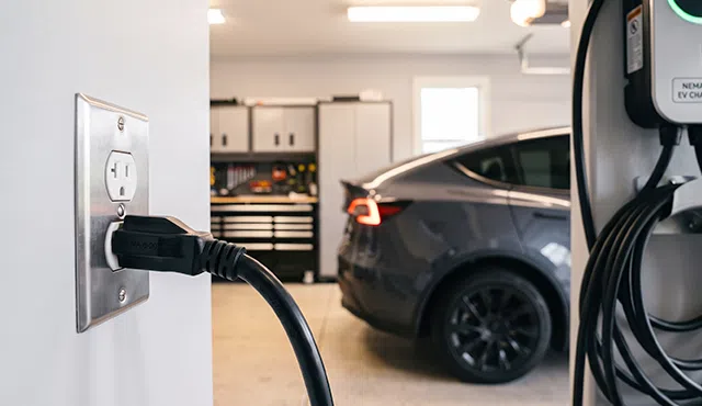 A close-up photograph of a NEMA 6-20 plug connected to a garage wall outlet for electric vehicle charging with an EV in the background